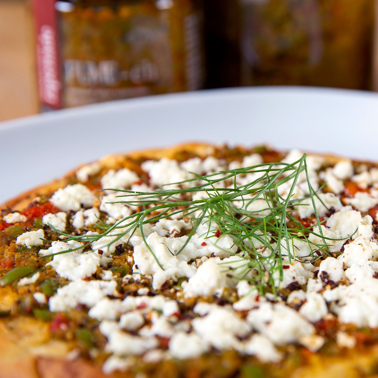 Pizza with crumbled cheese, herbs, and colorful toppings on a white plate; jars in the background.
