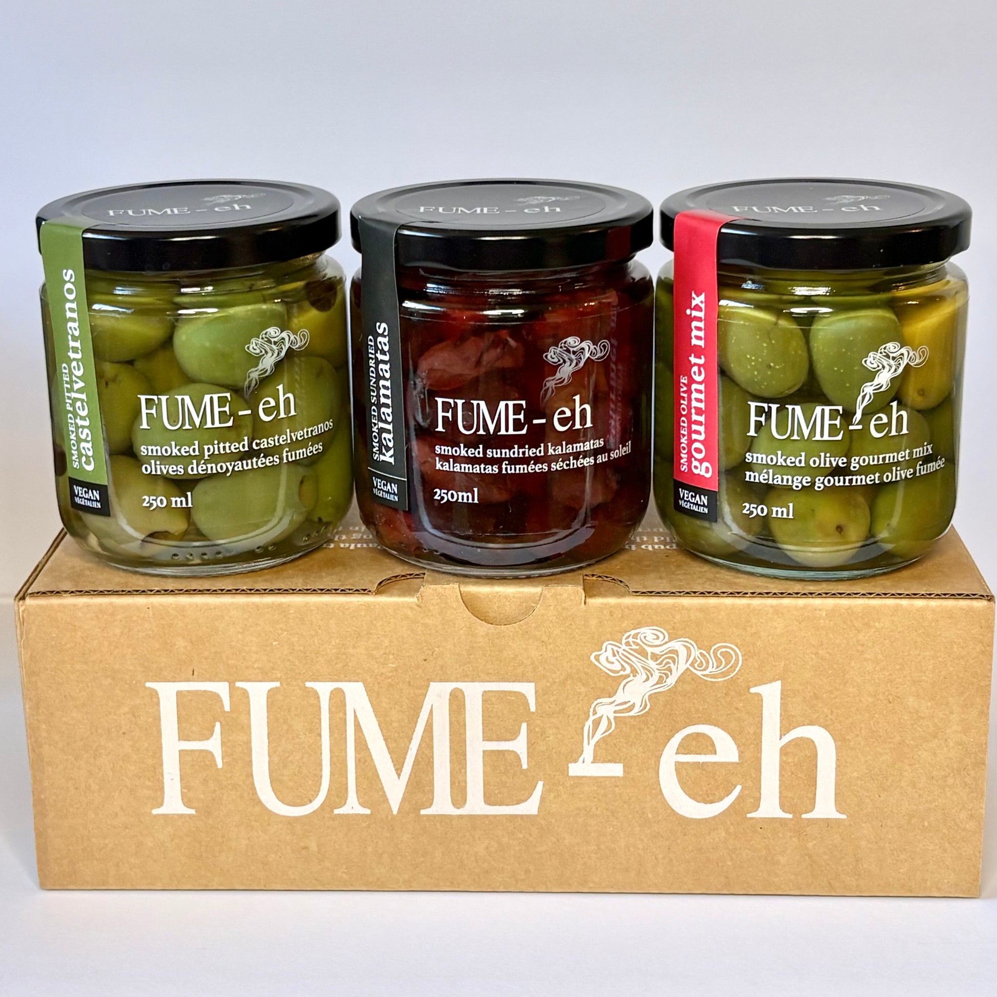 Three jars of FUME-eh olives on a brown box: green Castelvetranos, red Kalamatas, and gourmet mix.
