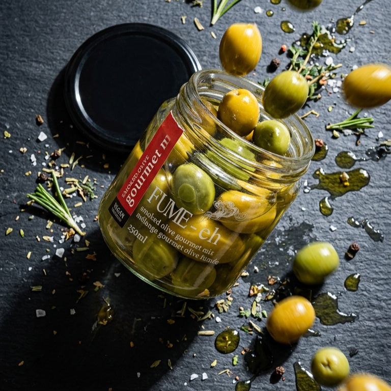 A glass jar of gourmet whole olives with a black lid placed beside it on a dark slate surface, surrounded by scattered olives, herbs, and oil droplets. The jar label reads 'Fume eh' and Smoked Olives Gourmet Mix