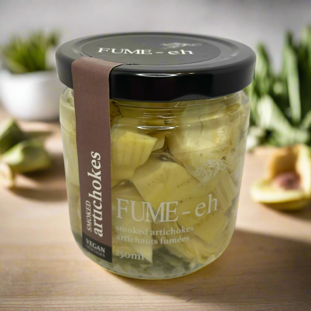 Jar of smoked artichokes with black lid and brown label on a wooden surface; blurred greens behind.